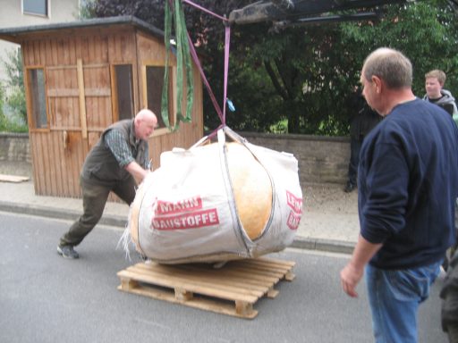 Ein Riesenkürbis, der in einem Big Bag an Gurten mit Hilfe eines Frontladers auf eine Palette gesetzt wird