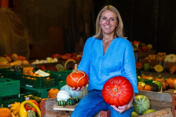 Frau in einer Scheune voller Kürbisse, sie hält zwei rote Exemplare in jeweils einer Hand